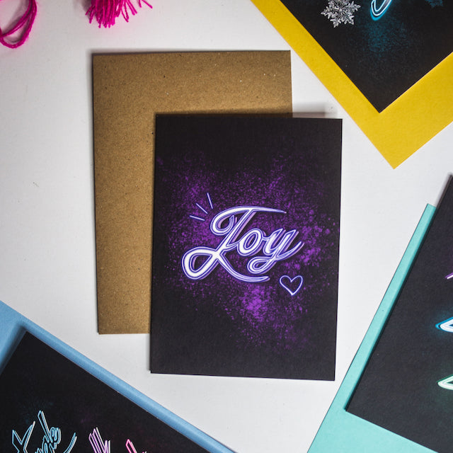 Purple Joy neon writing in purple splatter on black background christmas card by wayward