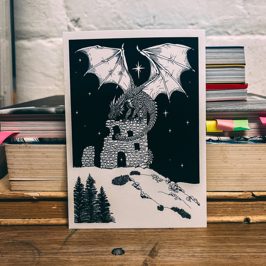Illustration of a dragon over a castle on a black background, placed on a wooden surface with books in the background.