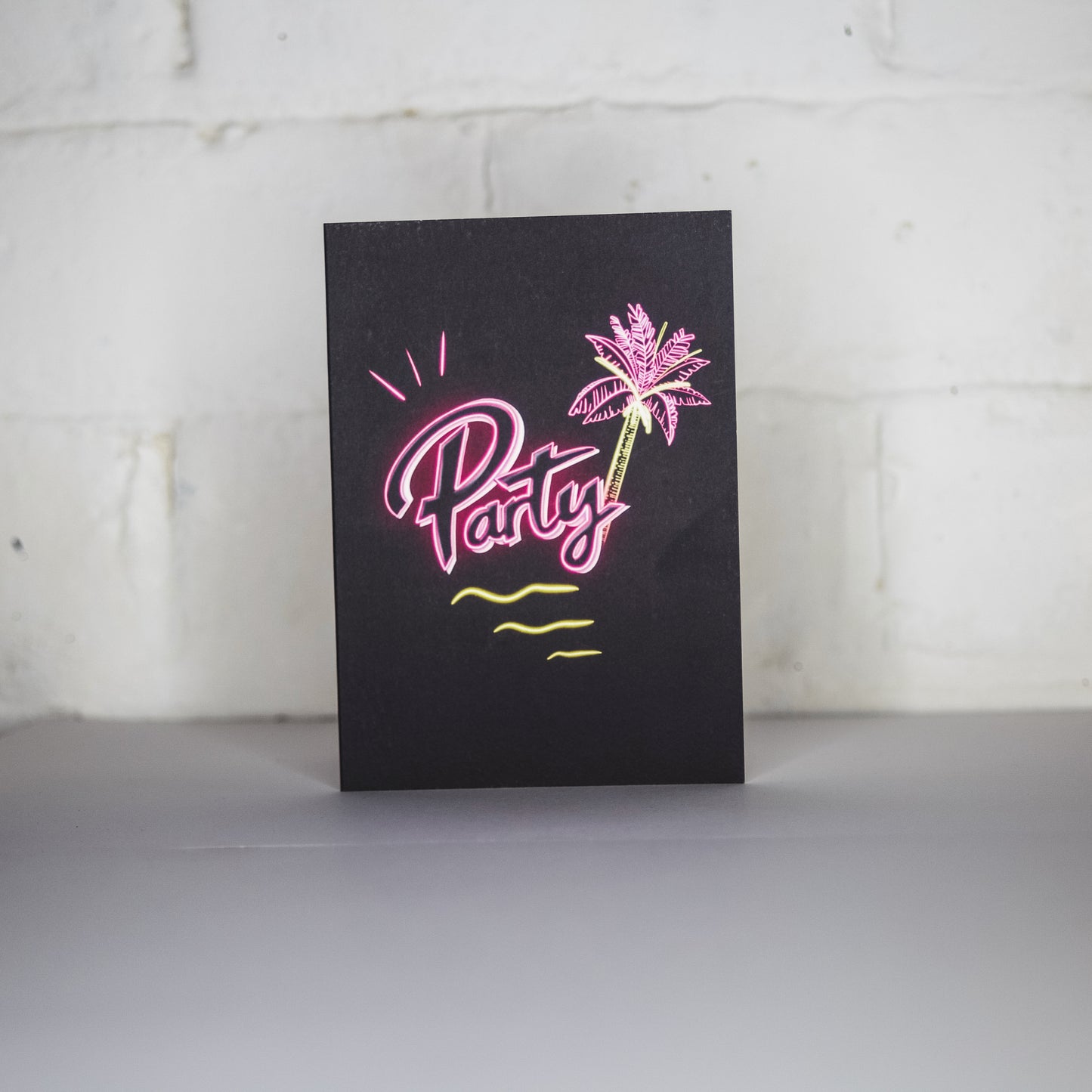 Black greetings card with party written in pink neon with a palm tree by wayward