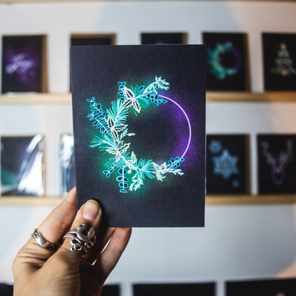 Christmas card by Wayward. Glowing green and purple neon wreath on black background being held